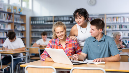 Obraz premium Three schoolchildren preparing for lessons in the school library are looking for information on a laptop and taking abstracts in a copybook