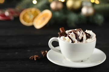 Tasty hot cocoa drink with whipped cream in cup and spices on black wooden table, space for text