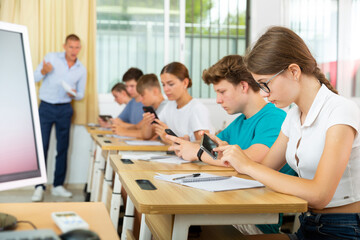 Fototapeta premium Concentrated teenage girl with group of fellow students sitting at lesson in classroom, using mobile phone