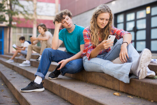 Teenager girl sitting on steps near school building and using her smartphone. Her freind sitting nex to her and looking at her.