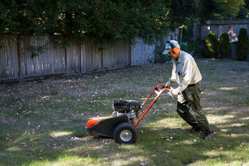 Senior man operating a gas powered stump grinding machine to cut back tree roots in a lawn, fall yard maintenance

