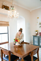 A woman smiling while carefully placing a vase of flowers on a wooden dining table under a chandelier.