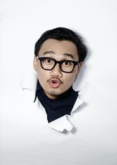 Happy asian man breaks through paper wall has fun looks gladfully at camera wears spectacles,...