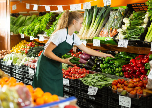Focused fifteen-year-old girl who works part-time in a store as a trainee saleswoman puts green pepper on showcase