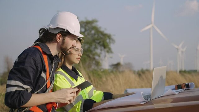 Renewable energy engineers working on wind turbine projects. Technician workers using tablet and laptop work outdoors at windmill farms. Sustainable renewable energy and environmentally friendly. 
