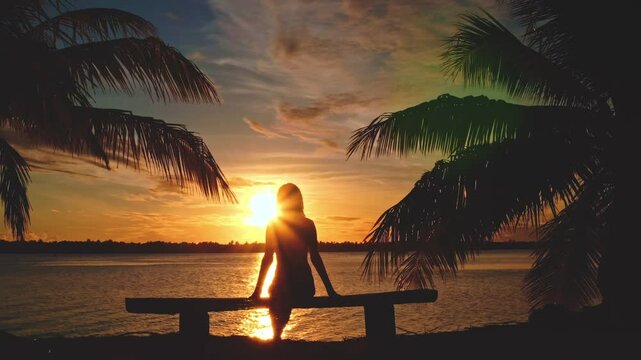 Woman sitting on a bench between palm trees, gazing at a stunning sunset on a tropical island in french polynesia's maupiti