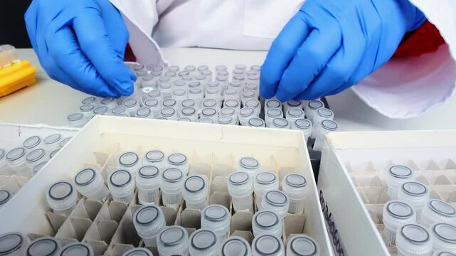 ab technician wearing gloves handles patient samples in test tubes in a genetic laboratory. The tubes are neatly arranged in a tray, indicating precise work with genetic material or blood diagnostics
