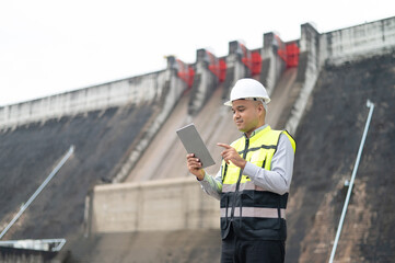 Smiling young Asian civil engineer standing with his arms crossed wearing a helmet and using a walkie-talkie. View and inspect the operation of the dam's sluice gates using the Quality Monitor tablet.