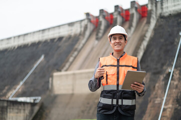 Smiling young Asian civil engineer standing with his arms crossed wearing a helmet and using a walkie-talkie. View and inspect the operation of the dam's sluice gates using the Quality Monitor tablet.