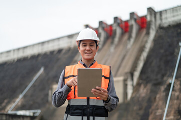 Smiling young Asian civil engineer standing with his arms crossed wearing a helmet and using a walkie-talkie. View and inspect the operation of the dam's sluice gates using the Quality Monitor tablet.