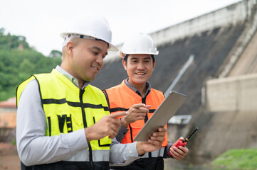 Smiling young Asian civil engineer standing with his arms crossed wearing a helmet and using a walkie-talkie. View and inspect the operation of the dam's sluice gates using the Quality Monitor tablet.