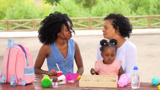 African grandmother and mother playing with girl in a park using toys sitting in a picnic table
