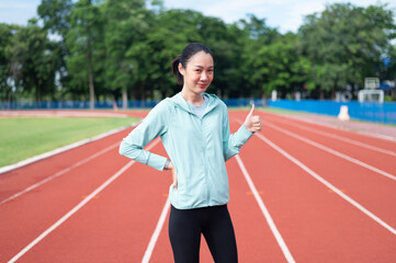 Fototapeta premium Asian woman wearing blue sportswear Stand in a pose with your hands showing the excellent sign. outdoor exercise Exercise in the stadium She is exercising and holding a water bottle. active living