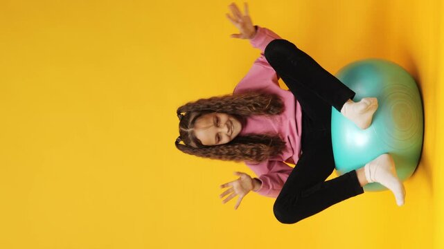 Vertical video. Joyful child with curly hair bouncing on fit ball wearing sweatshirt smiling while doing fun exercises enjoying playful activity and carefree fitness isolated over yellow background