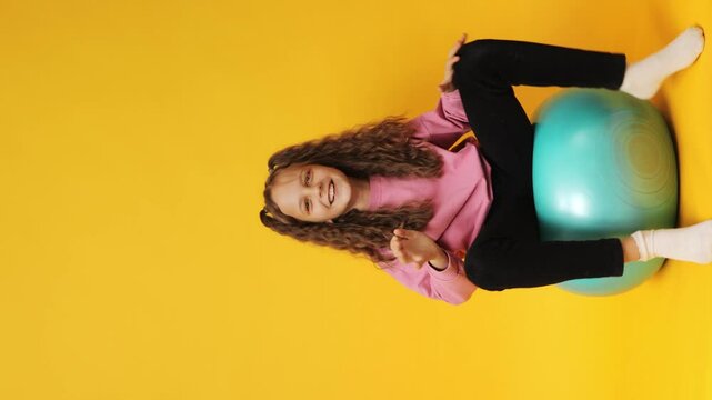 Vertical video. Happy young girl playing with gymnastic ball smiling joyfully jumping and balancing having fun during indoor activities isolated over yellow background