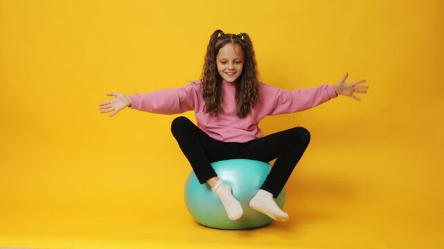 Adorable female kid with makeup bouncing on fitness ball enjoying playful and active lifestyle engaging in fun gymnastic exercises feeling carefree isolated over yellow background