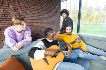 A group of friends is relaxing on a sofa in a cozy, modern living room, while one of them plays an acoustic guitar