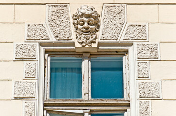  A close-up of a window on a building facade, featuring a grotesque mask carved into the stone above it. The window has a simple wooden frame and is partially open.