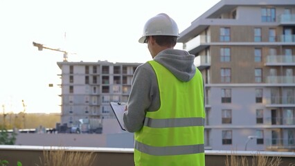 Man engineer with a white protective helmet and safety vest is writing on a clipboard while inspecting a construction site in early morning at sunrise. Architect concept