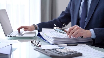 Businessman using a laptop computer and magnifying glass while analyzing financial documents at...