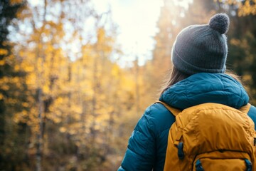 Fototapeta premium Adventurer among an Autumn Landscape. Back View of Young Woman Wearing Knitted Hat, Jacket, Backpack in Forest Woods Nature. Beauty and Adventure. Vibrant colorful fall scenery. Travel hike. Journey