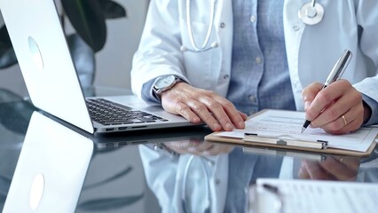 Doctor man wearing whit medical coat over a blue shirt is writing medical records on a glass desk...