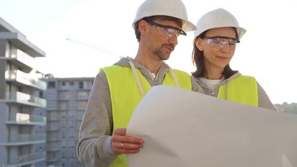 Architects or engineers wearing safety helmets and vests holding blueprint and discussing something on construction site at sunrise, front view. Architecture and teamwork concept