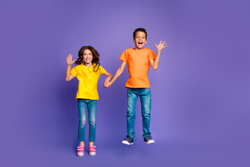 Full length photo of lovely two little kids jump wave hand dressed stylish orange yellow garment isolated on purple color background