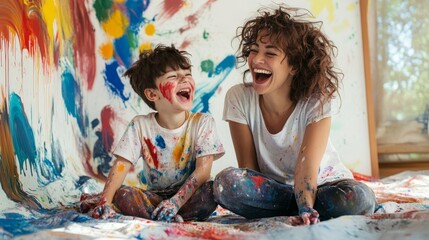 A joyful mother and son share a playful moment, covered in paint, surrounded by vibrant colors on a wall and floor, capturing a fun artistic experience together.