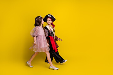 Full size profile photo of two little kids hold arms walk empty space halloween pirate princess costume isolated on yellow color background
