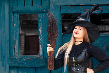 A fat witch in a black corset and hat holds a broom against the background of an old hut