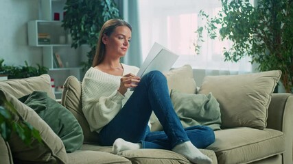 Woman sits comfortably on a sofa in her living room, leisurely reading through documents. The scene captures a peaceful atmosphere filled with natural light and tranquility