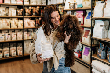 Beautiful young woman enjoying in modern pet shop together with her adorable brown toy poodle.