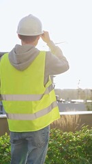 Man engineer with white helmet and safety vest is taking notes on a clipboard while inspecting a construction site at sunrise, back vertical view