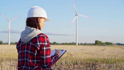 Female engineer wearing a white protective helmet is taking notes with a clipboard in a field with wind turbines, as the sun sets. Clean energy concept