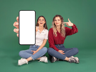 Full body two young caucasian women sit floor legs crossed show empty blank screen mobile phone mock up close up to camera pointing finger to touch screen isolated green background, copy space.