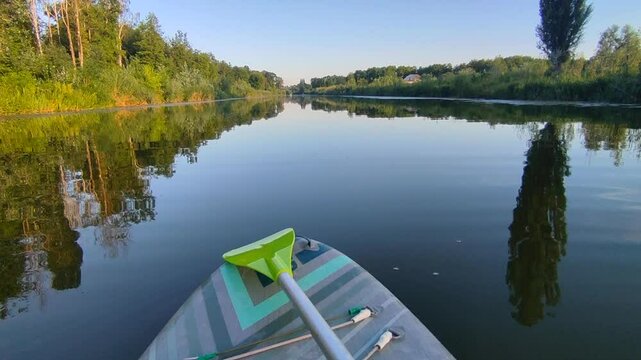 SUP board with green paddle on surface of river water with trees on banks on calm windless summer evening. Person swimming on SUP board on pond with reflective water surface. Tourism travel rest sport