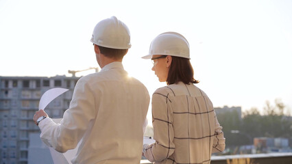 Man and woman architects, wearing safety helmets and glasses, are examining blueprints at a construction site during a sunset, back view. Architecture concept