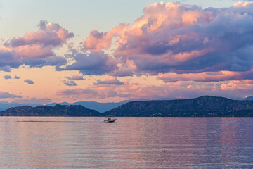 Sunset view of the Corinthian Gulf with pink clouds reflecting on calm waters, a speedboat in the distance, and rugged mountains in the background. Corinth, Greece