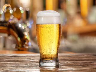 Glass of chilled beer on wooden bar table top and blurred bar interior at the background.