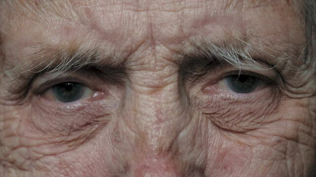 Close up of face of old elderly woman granny female with large wrinkled face of advanced age. Sad thoughtfully looks at camera, illness from nervous tic with tremor, twitches the eyelid of eye.