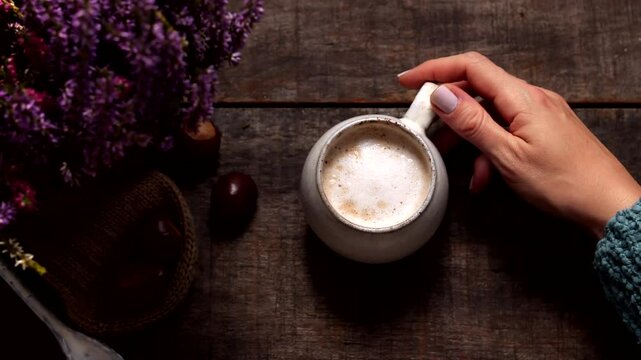 hand holds a mug filled with creamy froth coffee on a rustic wooden table. Warm comfort and relaxation. Dried lavender flowers, scattered chestnuts. Cozy, autumnal atmosphere. Warm cappuccino.