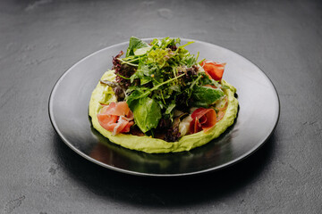 Fresh salad with avocado cream and prosciutto, served on a black plate, perfect for gourmet dining