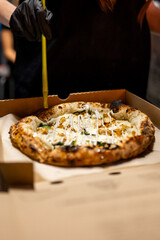 A freshly baked pizza with various toppings in an open cardboard box, held by a person wearing a...