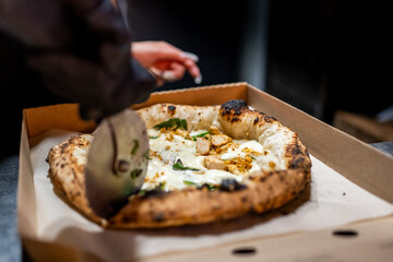Close-up of a hand slicing a hot, fresh pizza with mozzarella and basil in a cardboard box. Steam rises, highlighting the texture of the crust and the freshness of the ingredients