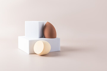 Two cosmetic egg-shaped blender sponges on a white square catwalk. Beige background. Front view. Care and beauty concept. Makeup tool.