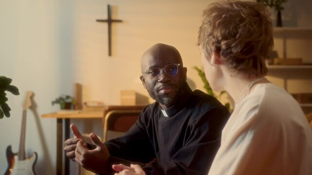 African American priest wearing cassock and glasses sitting on couch in living room and talking to Caucasian woman while visiting her at home