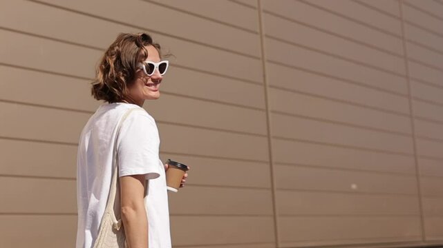Young woman holds take away eco paper cup with recycle lid hot cold drink, coffee or tea, has fun rotating outdoors on modern building wall backdrop. Beverage to go on sunny day. Zero waste lifestyle