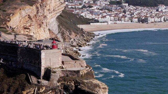 Aerial drone approaching the stunning cliffs and famous lighthouse at Nazare, Portugal during a sunny day at the beach with the village of Nazare in the background. Nazare is famous for giant waves