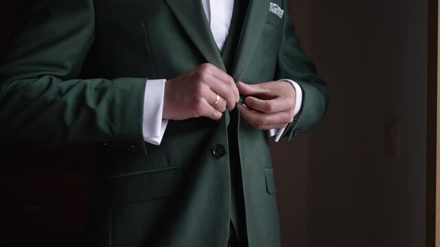 Man buttoning his green suit jacket, wearing a wedding ring and cufflinks in a formal setting